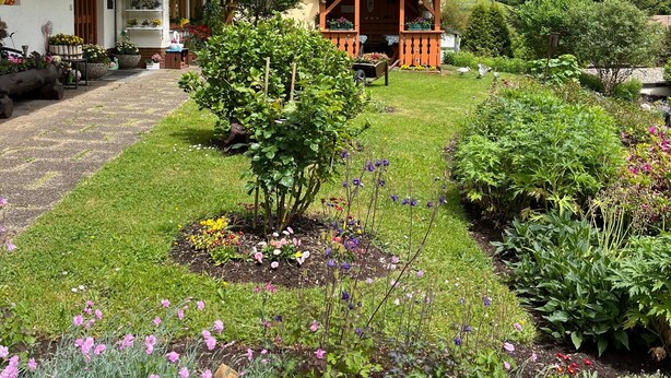 Um chalé tradicional com varanda de madeira, um jardim com diversas flores e montanhas ao fundo.