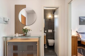 Un baño moderno con un lavabo de vidrio, un espejo redondo, un revestimiento de pared de madera, un inodoro y una cama con una mesita de noche.