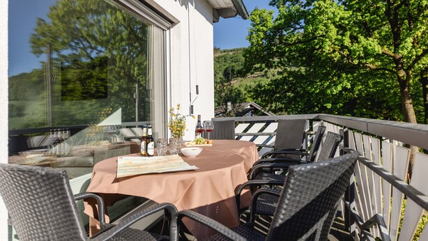 Un espace de restauration en plein air avec une table dressée pour deux personnes, entouré de chaises en osier, d’une rambarde en verre et offrant une vue sur une colline.