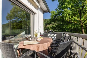 Ein Essbereich im Freien mit einem Tisch für zwei Personen, umgeben von Korbstühlen, einer Glasreling und Blick auf einen Hügel.