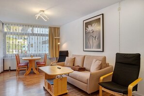 Una sala de estar con sofá, mesa y sillas. Hay un cuadro en la pared y una ventana con cortinas.