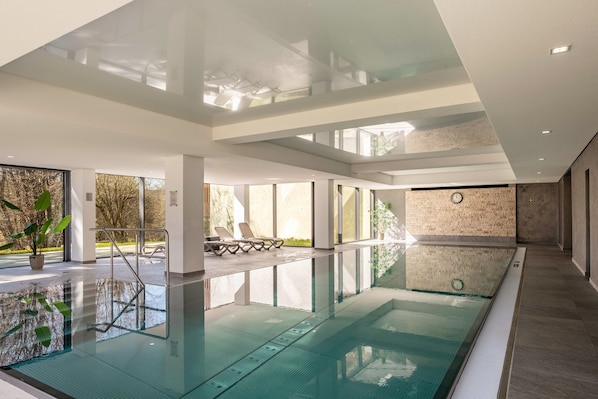 A modern indoor swimming pool with clear glass walls, a sunlit area with lounge chairs, and a stone wall with a clock.