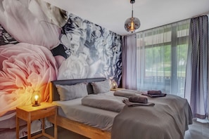 A bedroom with a large floral mural, a bed with grey bedding, a bedside table with a lamp, and a view of greenery through sheer curtains.