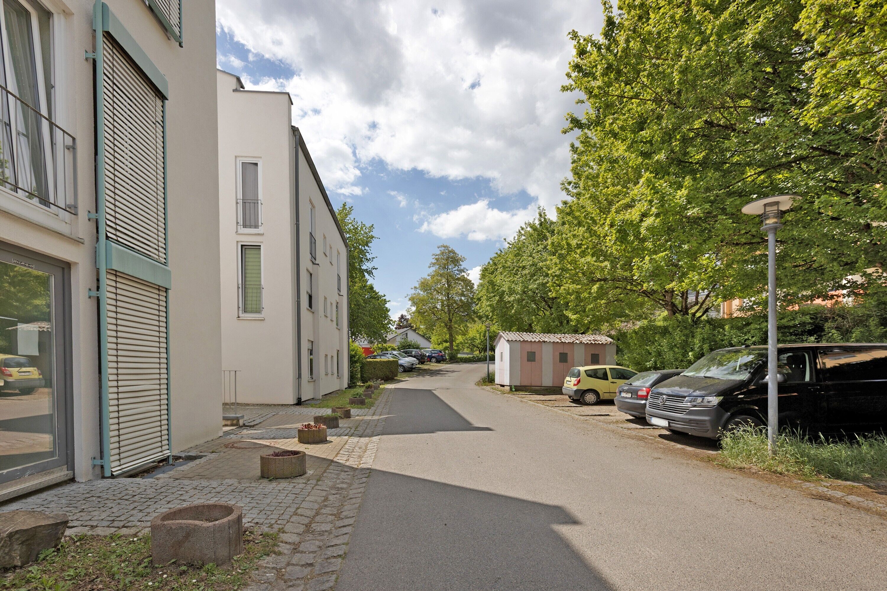 A residential street with parked cars, a small shed, and a building with a shuttered window.