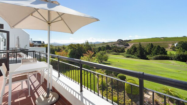 Un balcon avec une table et une chaise blanches, un grand parasol et une vue sur un paysage verdoyant.