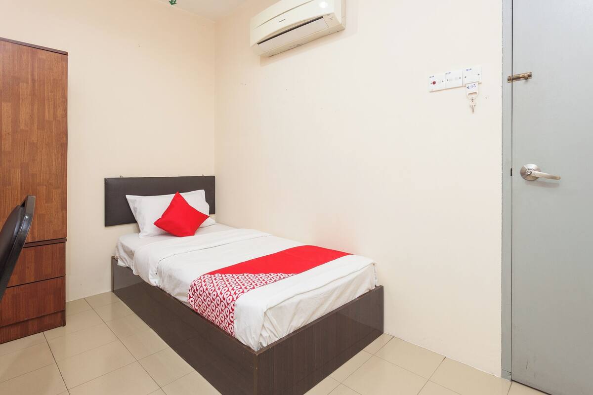 A single bed with a red and white bedspread, a wooden wardrobe, and a white air conditioning unit mounted on the wall.
