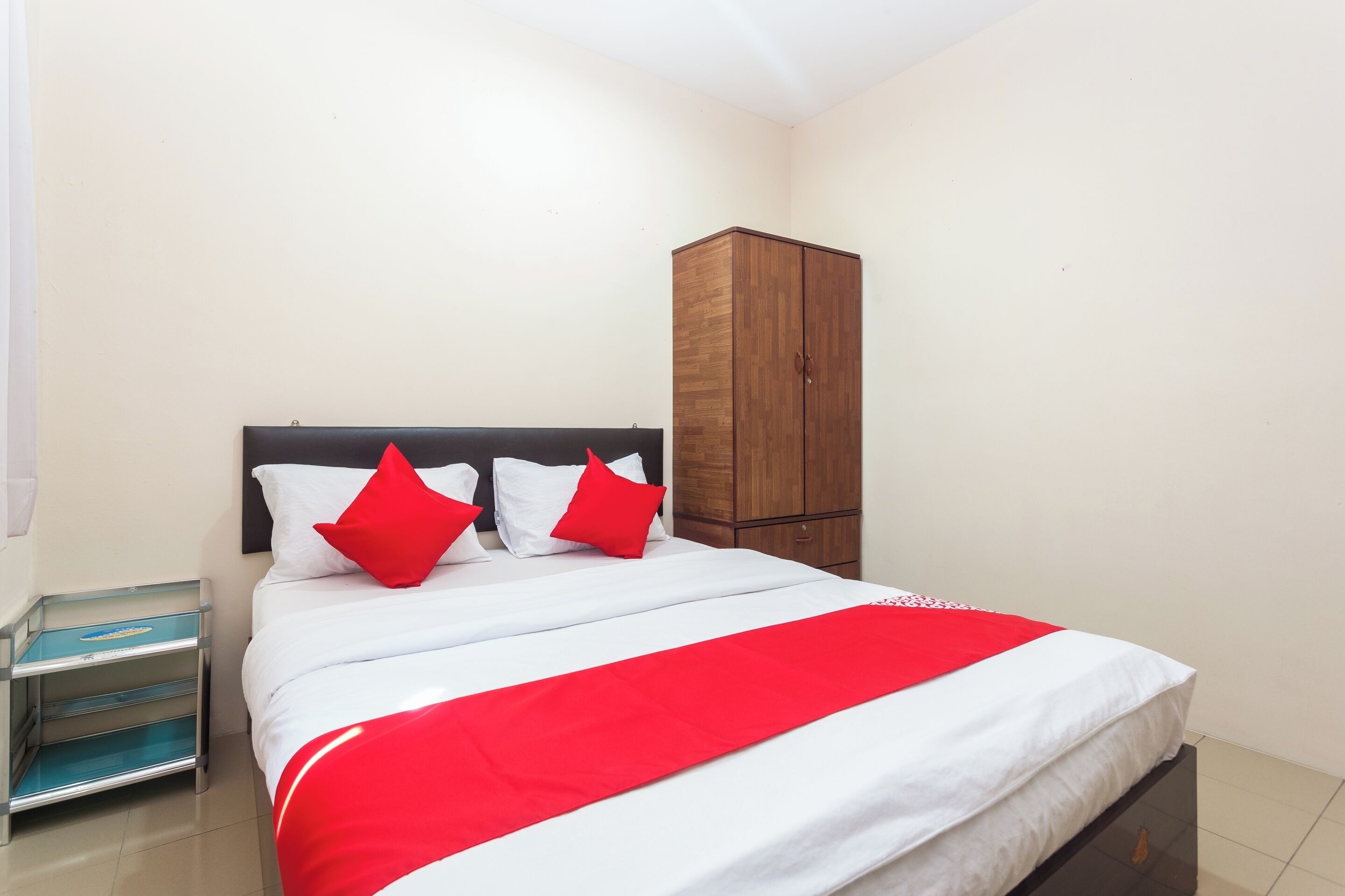 A neatly made bed with white and red bedding, a wooden wardrobe, and a small bedside table with a blue tray.