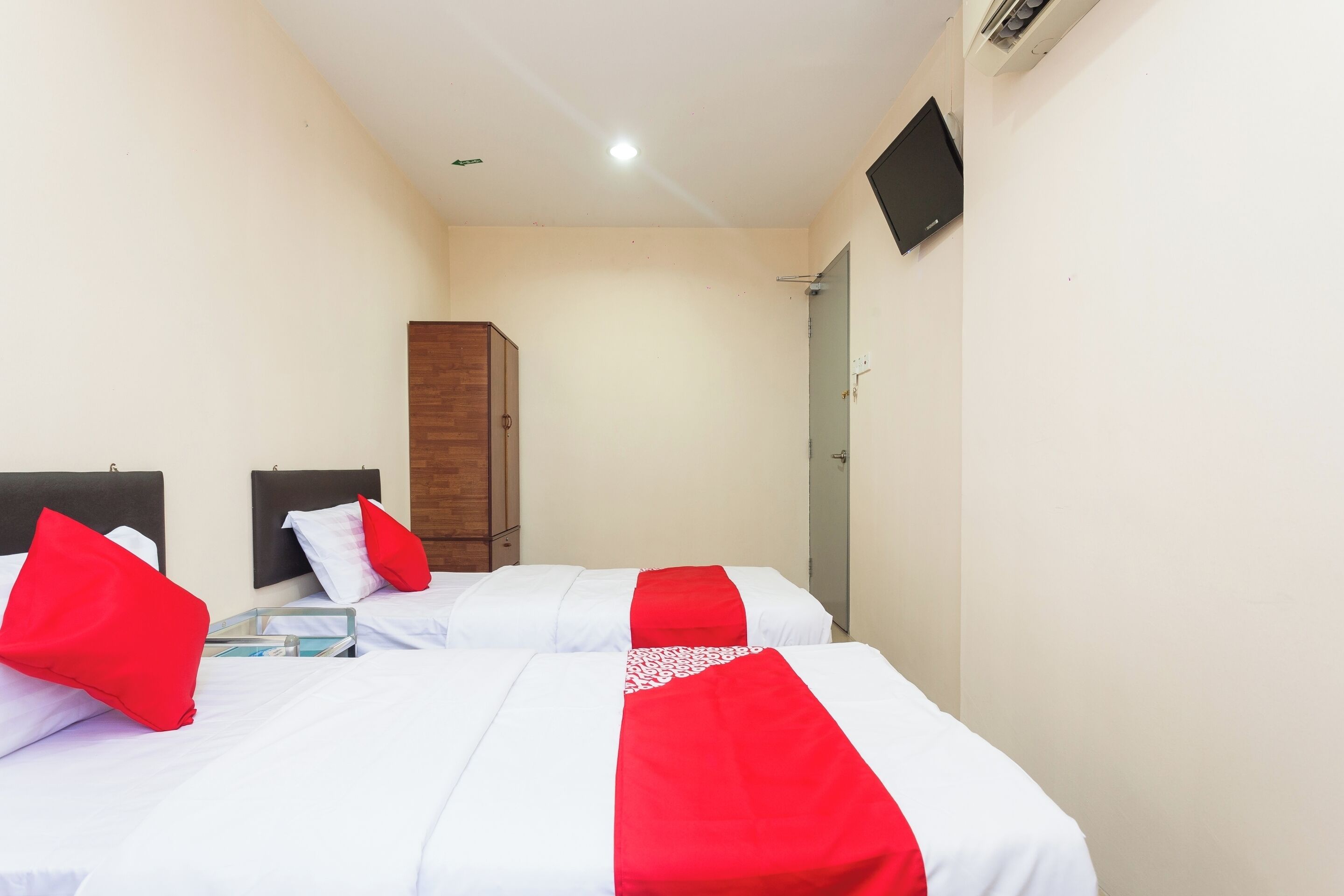 Une chambre d’hôtel avec deux lits, chacun agrémenté de draps blancs et de touches de couleur rouge, une armoire en bois, un téléviseur à écran plat et une porte.