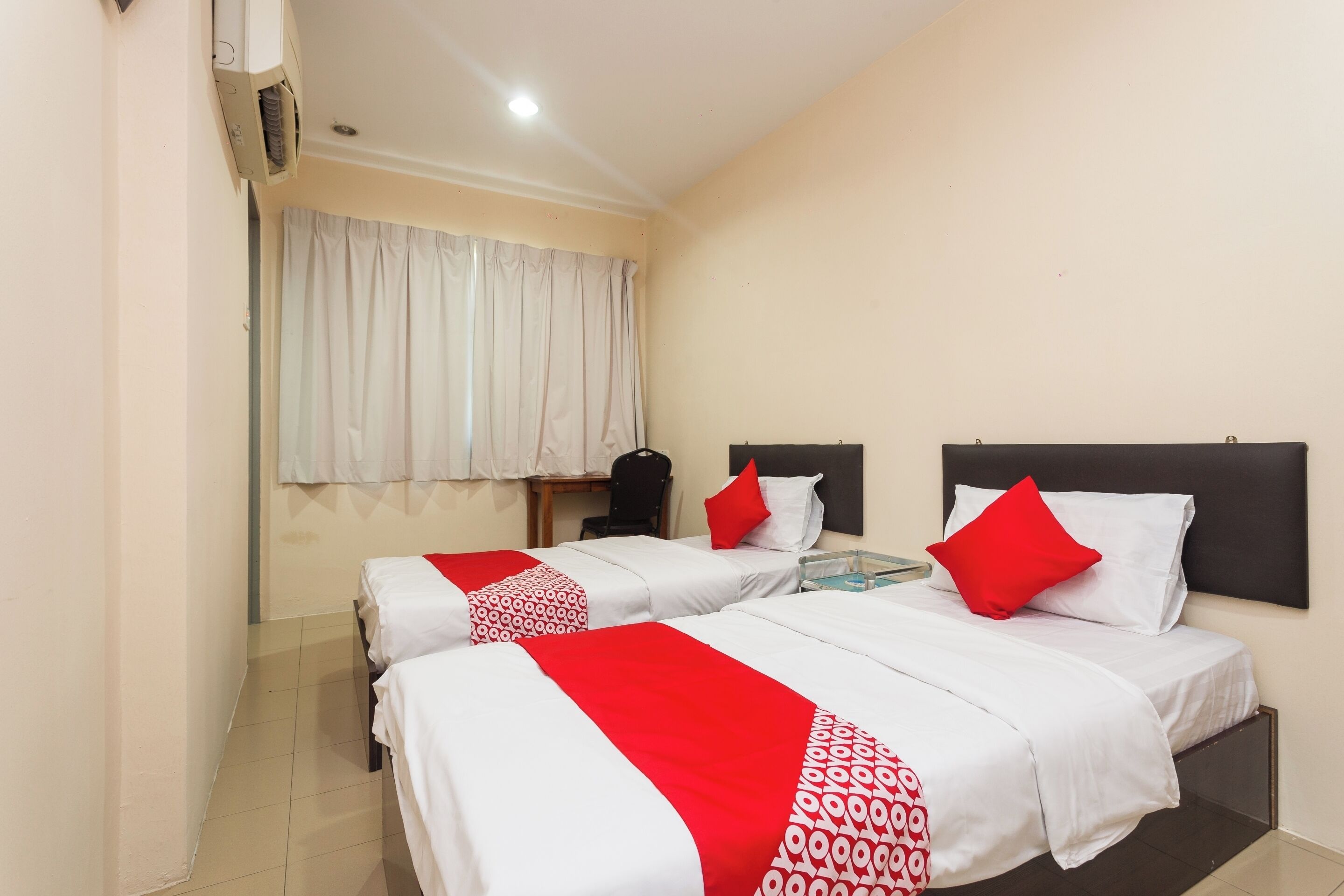 Habitación de hotel con dos camas, cada una con ropa blanca y detalles en rojo, una mesita y una ventana con cortinas blancas.