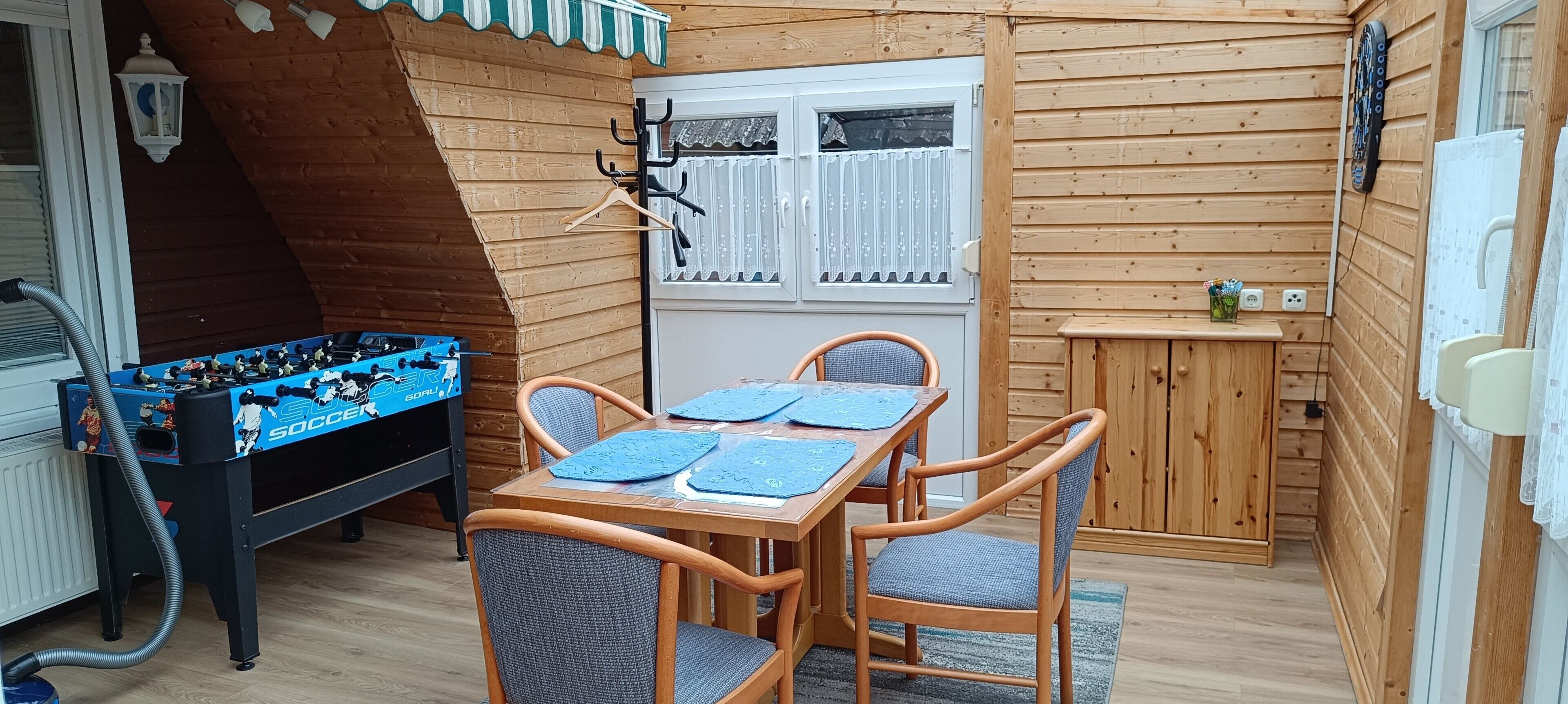 Ein Zimmer mit Holzboden, einem Kicker, einem Esstisch mit Stühlen, einem Fenster mit Vorhängen und einem kleinen Schrank.