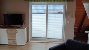 A room with a television on a wooden cabinet, a sliding glass door with blinds, a lamp, and a radiator. - Holiday Home 'Ferienhaus Stamer Am Großen Meer' with Shared Terrace and Private Garden (Bedekaspel)
