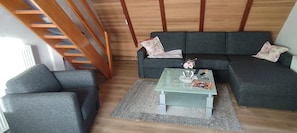 A living room with a grey sofa, a matching armchair, a small coffee table with a vase and remote control, and a wooden staircase.