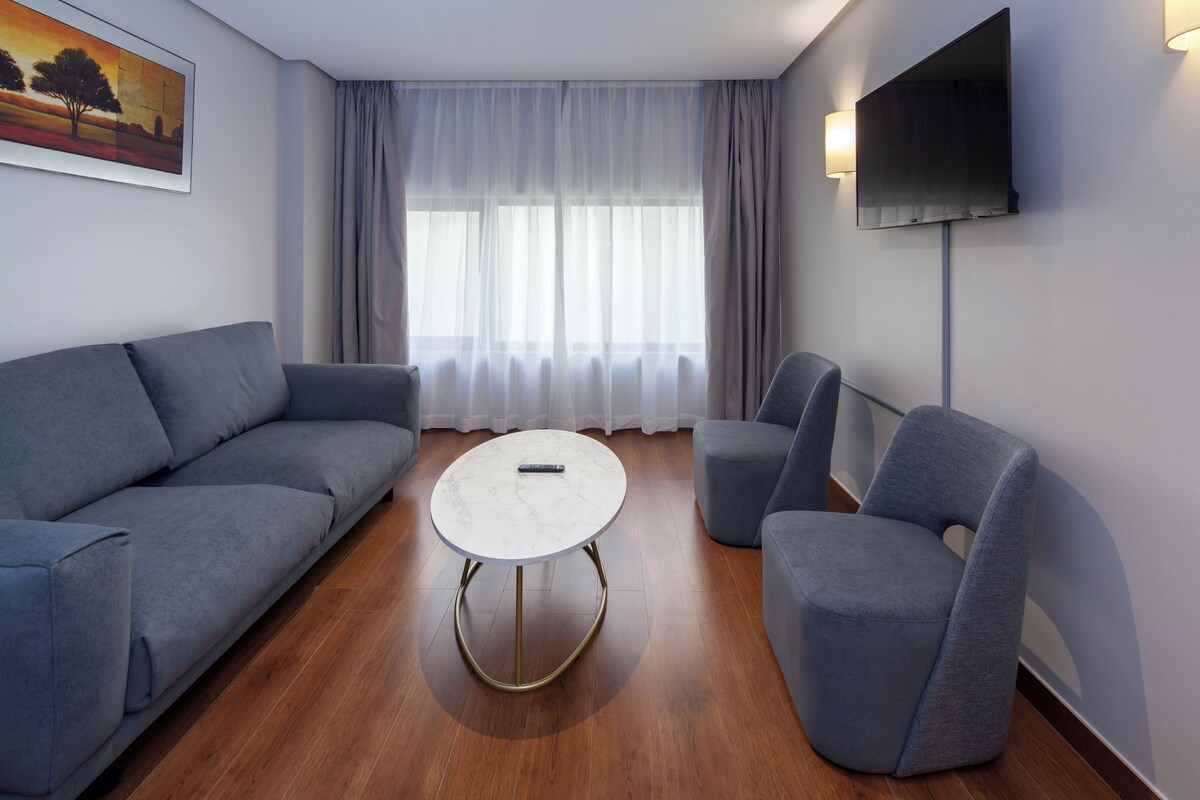 A modern living room with a grey sofa, a round coffee table, and two armchairs. A flat-screen TV is mounted on the wall, and a painting is visible on the wall.