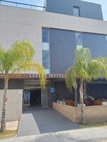 Exterior del Hotel Leflet con un diseño moderno, que presenta un techo plano, ventanas amplias y un área de entrada pavimentada con palmeras.