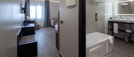 A modern hotel room with a wooden floor, a sitting area with a desk and chair, a large bed, a separate bathroom with a bathtub, and a large mirror.