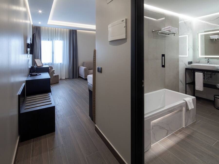 Une chambre d’hôtel moderne avec un plancher en bois, un coin salon comprenant un bureau et une chaise, un grand lit, une salle de bain séparée avec une baignoire et un grand miroir.