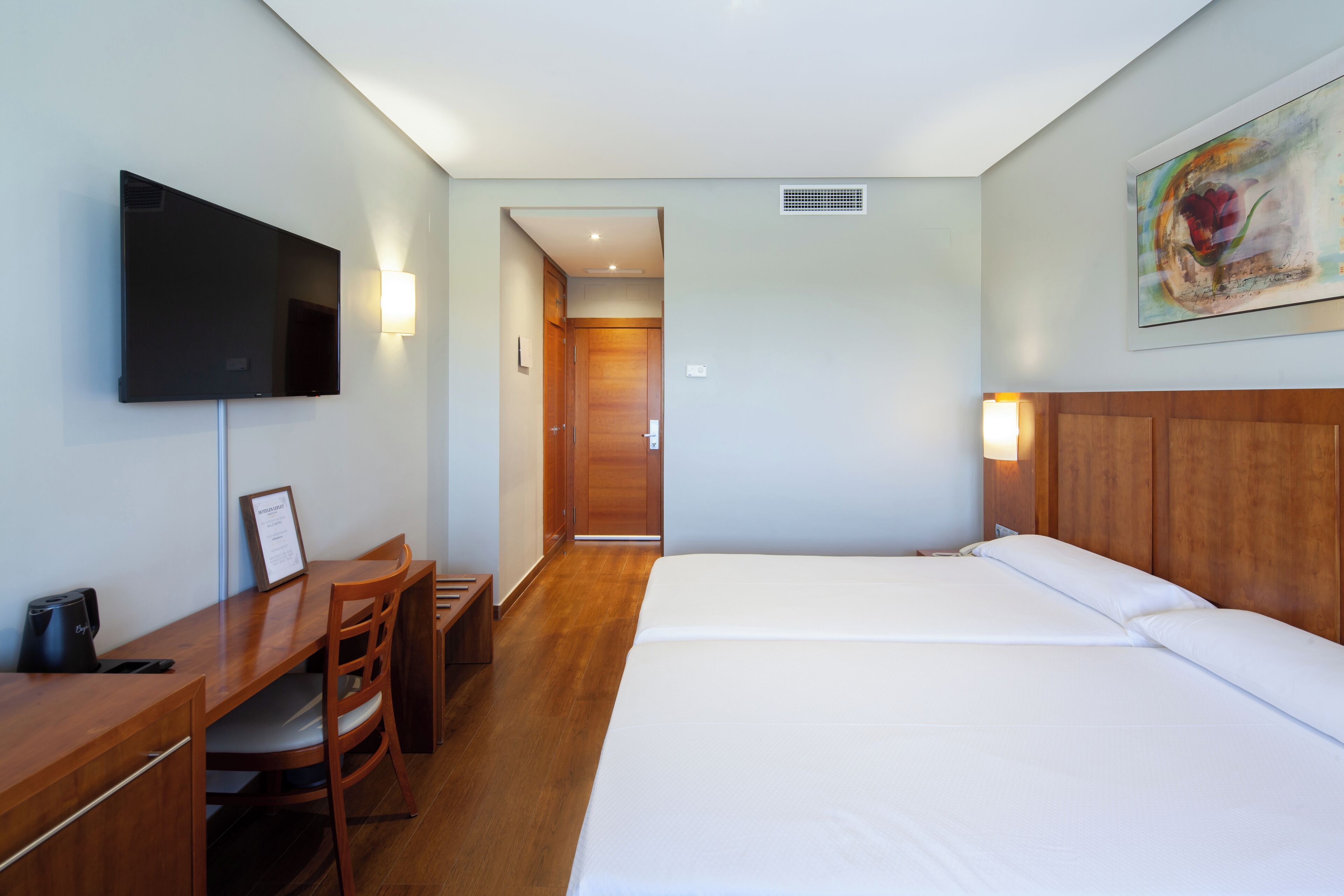 A hotel room with a bed, a desk with a chair, a flat-screen TV, and a painting on the wall.