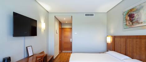 A hotel room with a bed, a desk with a chair, a flat-screen TV, and a painting on the wall.