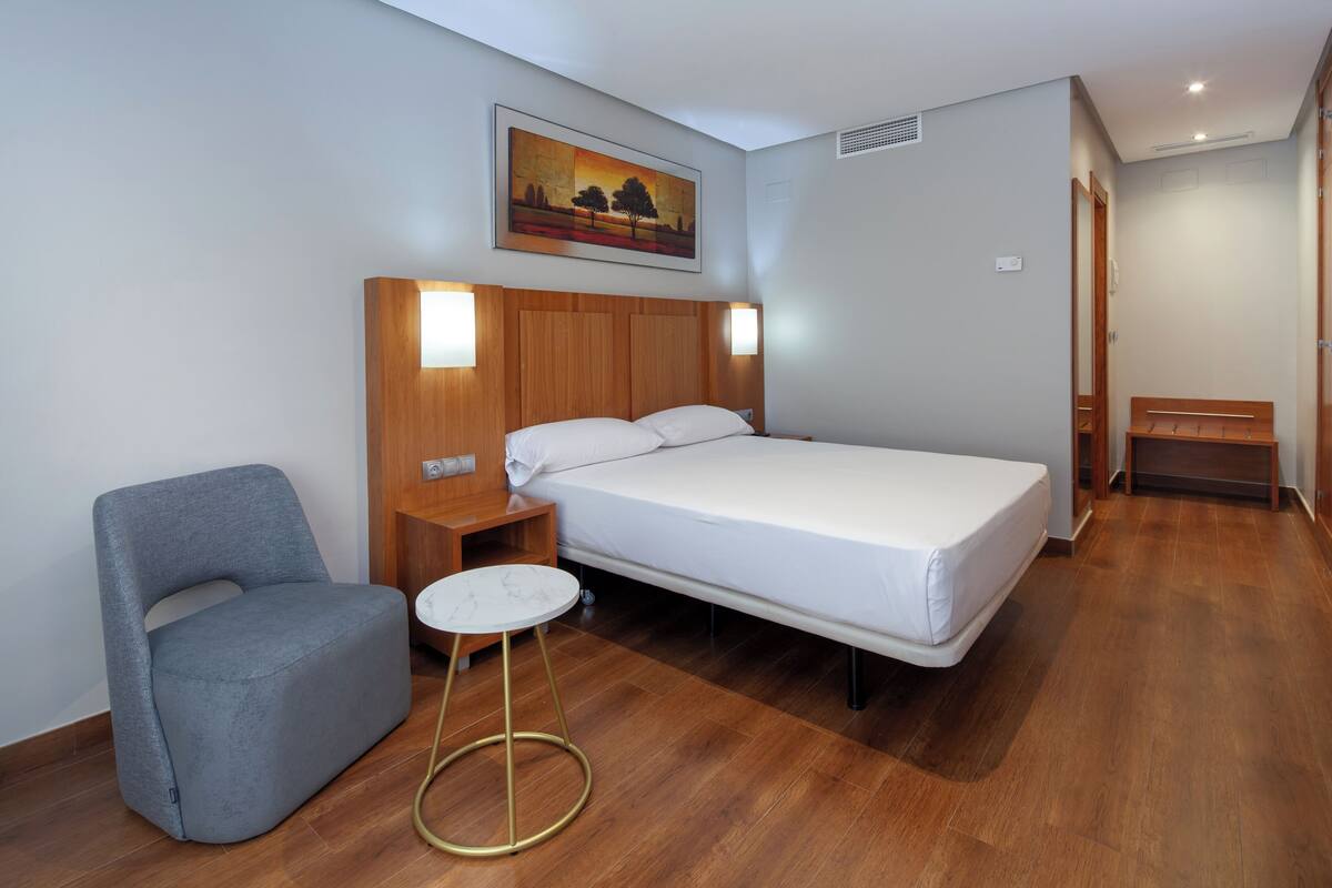 Una habitación de hotel con una cama, un sillón gris, una mesita redonda y un cuadro en la pared.