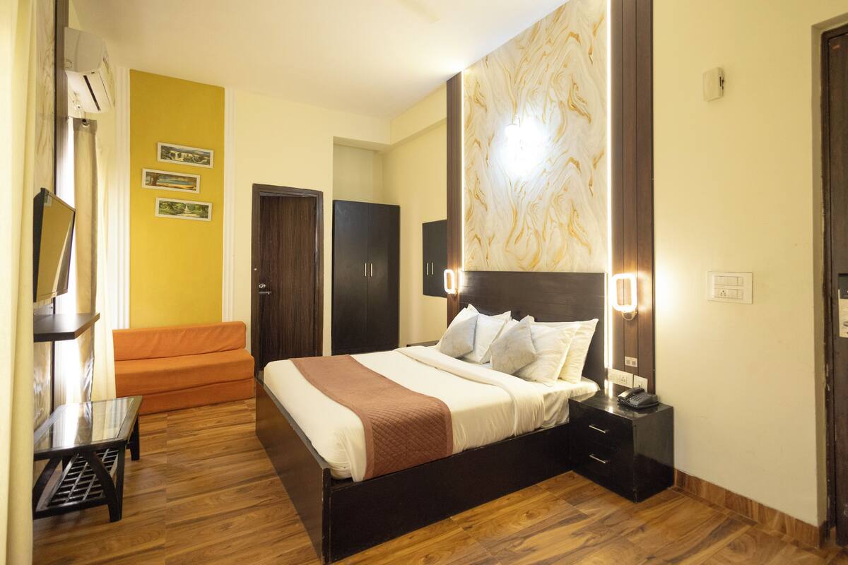 A hotel room with a wooden floor, a bed with white bedding, a nightstand, a desk, and a chair. There are two doors and a wall with a light fixture.
