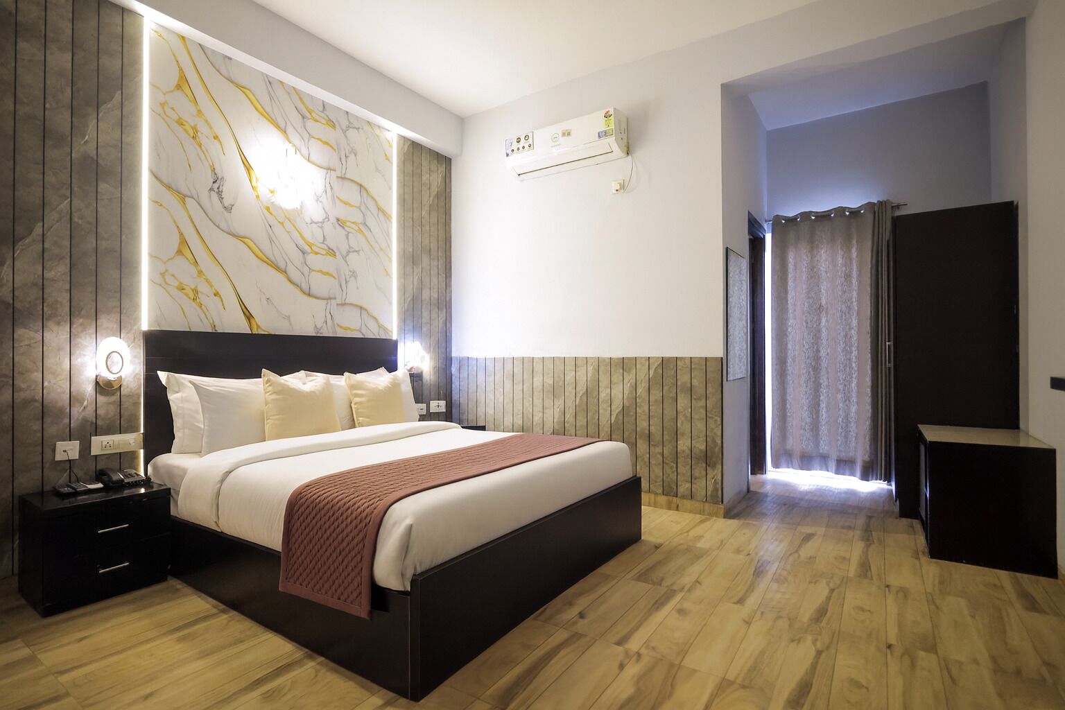 A modern hotel room with a large bed, wooden flooring, marble wall accents, and a built-in air conditioning unit.