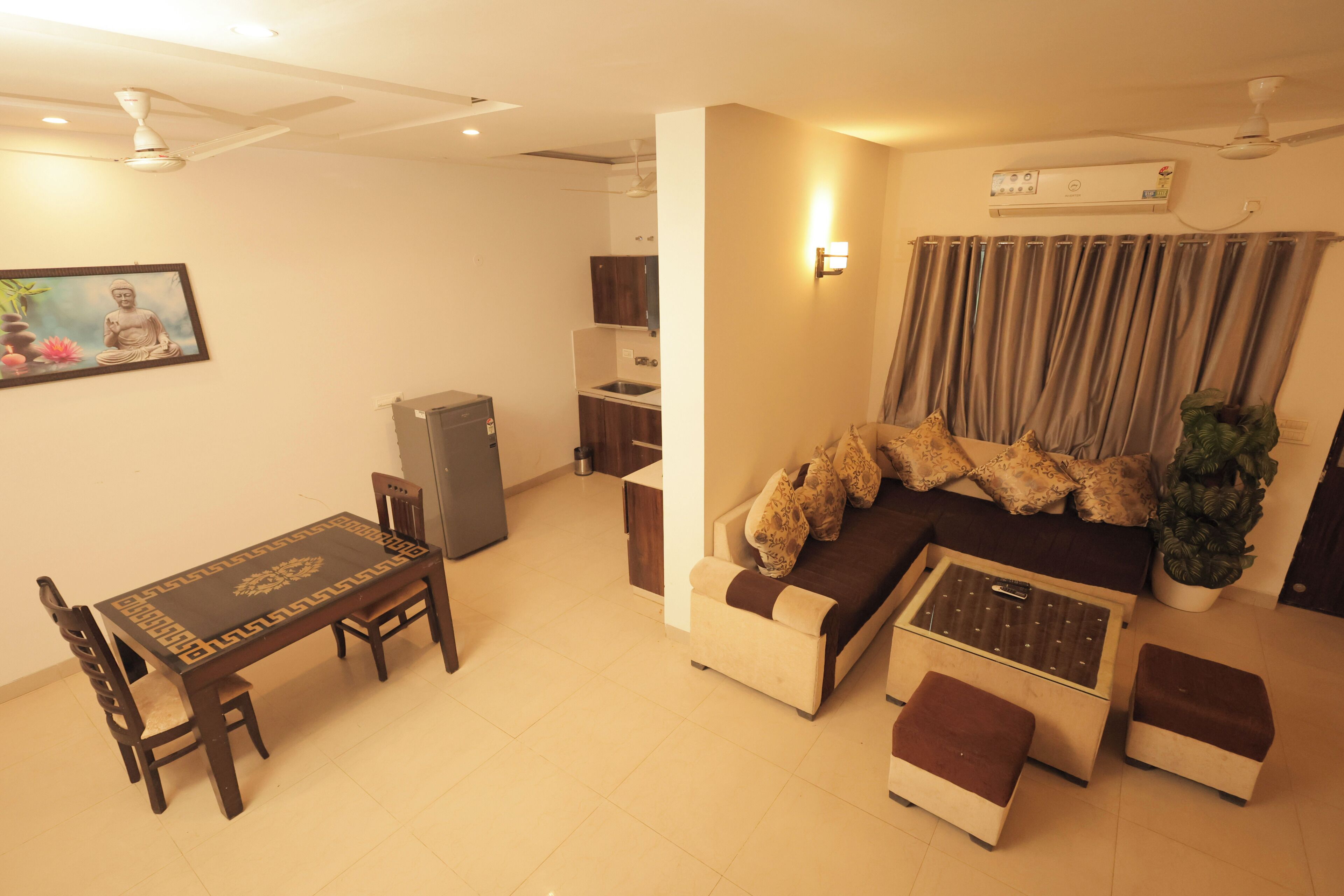 A living room with a sofa, ottomans, a dining table with chairs, a ceiling fan, an air conditioning unit, and a small kitchen area.