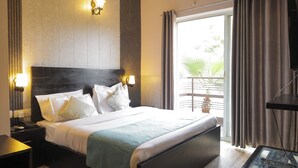 A hotel room with a large bed, bedside lamps, a ceiling fan, an air conditioning unit, a balcony with curtains, and a small table with a glass top.