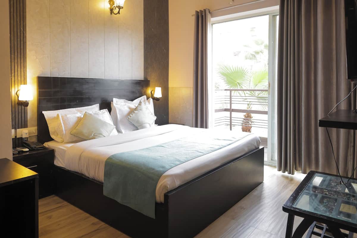 A hotel room with a large bed, bedside lamps, a ceiling fan, an air conditioning unit, a balcony with curtains, and a small table with a glass top.