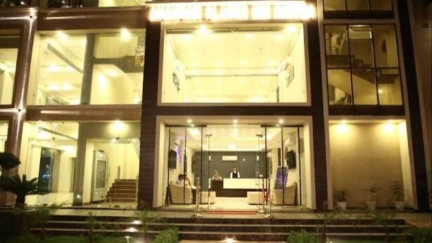 A modern hotel entrance with a reception desk, glass doors, and a staircase leading to the upper floor.