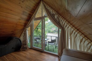 A room with a sloped ceiling, a large window with curtains, a wooden floor, and a small table with chairs. - Trishuli Villa Resort  (Benighat Rorang)