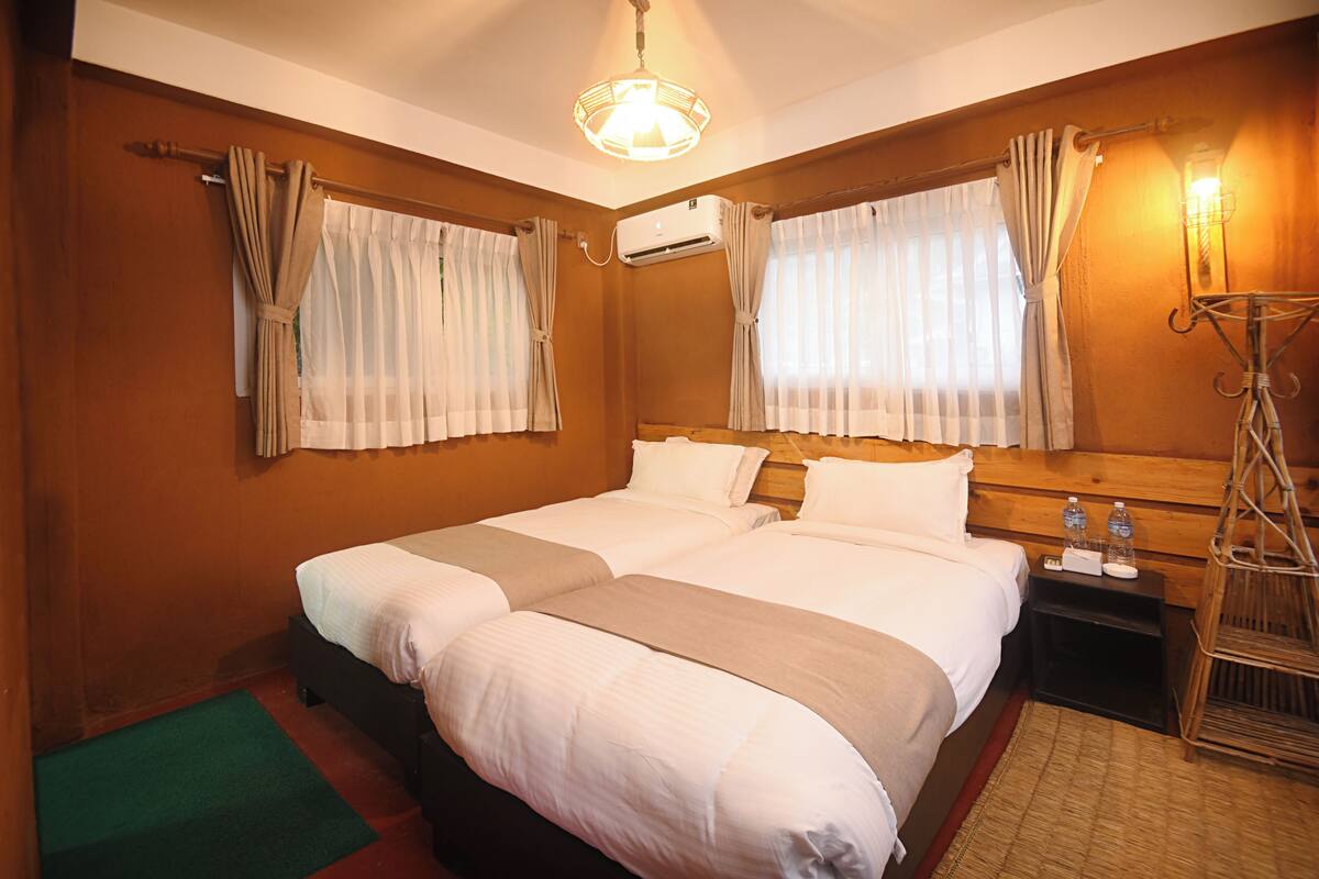 A hotel room with two beds, a wooden headboard, a bedside table with a lamp, and a green mat.