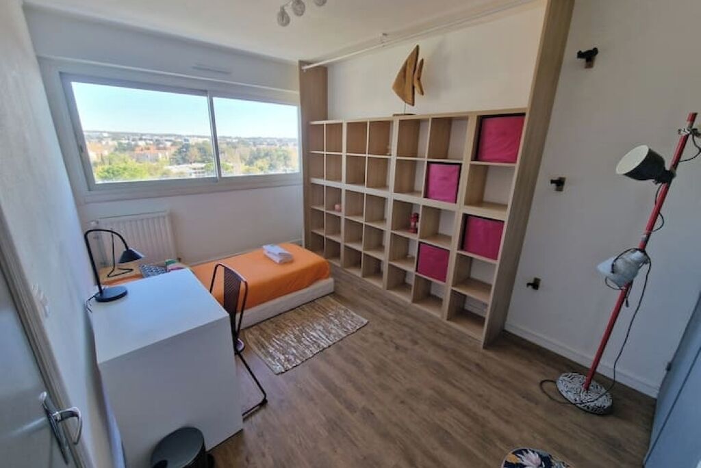 A compact room with a bed, a desk, a radiator, a window with a view, and a shelving unit with pink items.