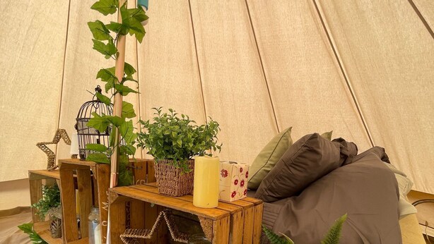 Et hyggeligt teltinteriør med en seng, en træhylde med planter og stearinlys samt en tæppe med et mønster.