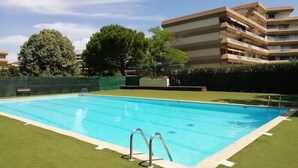 A rectangular swimming pool with two metal ladders, surrounded by well-maintained grass and a hedge. In the background, there are multi-story buildings with balconies.