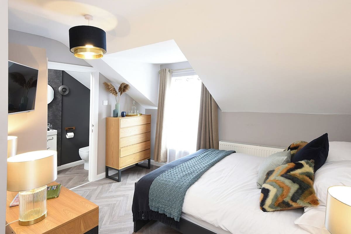 Un dormitorio moderno con cama, mesita de noche, lámpara, cómoda y vista al baño.