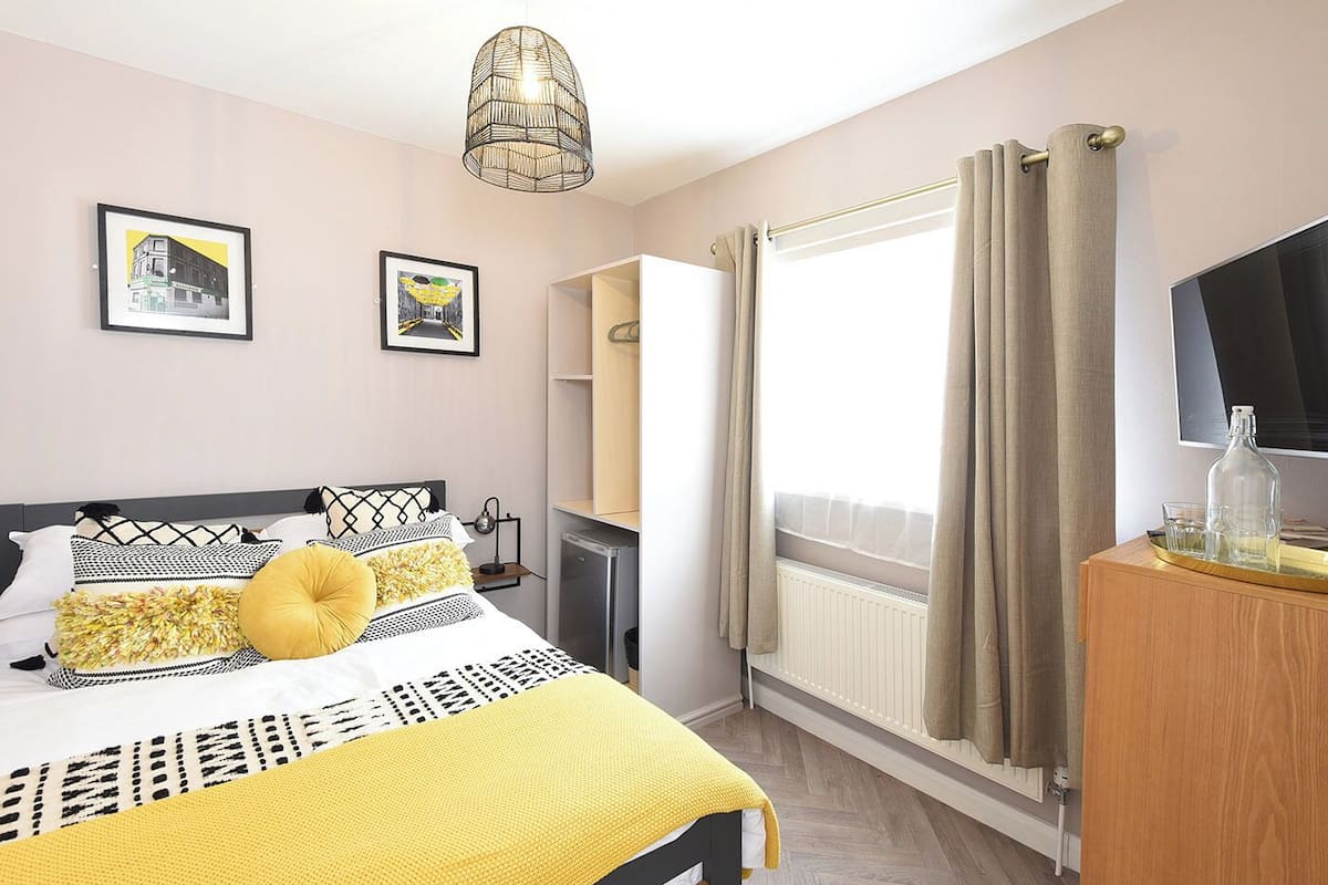Un dormitorio con cama, mesita de noche, televisor, radiador y ventana con cortinas.