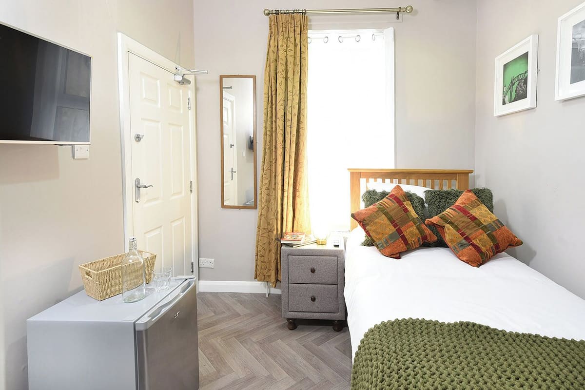 Un dormitorio con cama, mesita de noche, cómoda, espejo, ventana con cortinas y televisión montada en la pared.