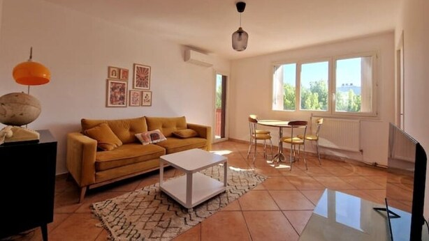 A living room with a sofa, coffee table, and dining table. There are framed pictures on the wall and a window providing natural light. - Large, modern, air-conditioned T4 (Montpellier)