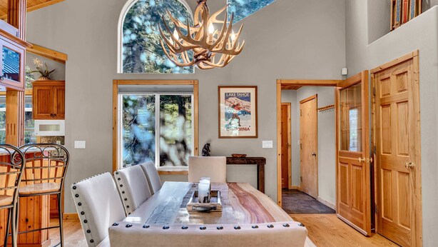 A spacious dining area with a long table, wooden chairs, and a chandelier. There are wooden cabinets and a large window with a view of trees. - Modern Retreat in Truckee with A/C & Game Room (Truckee)