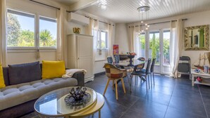 A spacious living area with a sofa, a glass coffee table, and a dining table with chairs. There is a large window providing natural light and a view of greenery outside. - Gorgeous home in Royan (Royan)