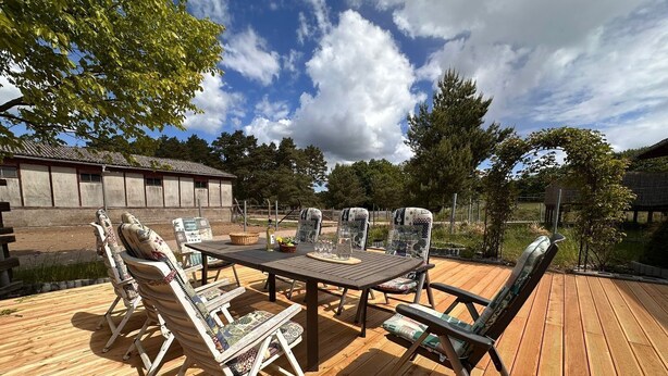 Een terras buiten met een houten tafel en stoelen, omgeven door bomen en een gebouw op de achtergrond.
