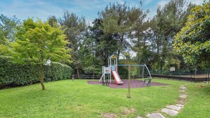 A playground with a slide and swings, surrounded by grass and trees.