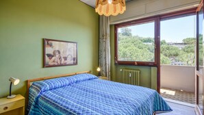 A bedroom with a bed, a nightstand, a lamp, a window with a view of trees, and a painting on the wall.