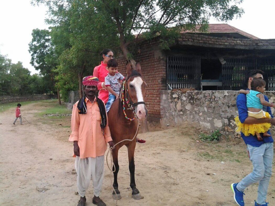 Un hombre conduce un caballo con un niño a lomo, otro niño caminando al fondo, y un edificio de ladrillo a lo lejos.