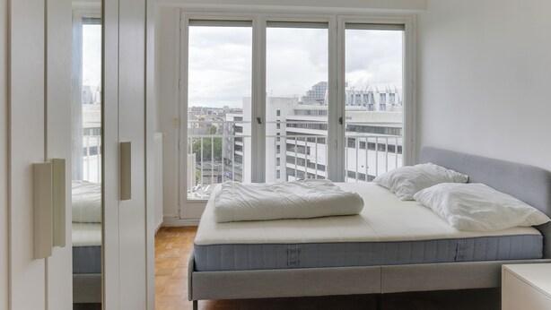Een slaapkamer met een bed, uitzicht op de stad door grote ramen en een deur naar een balkon.