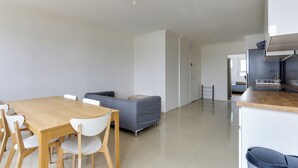 Een moderne, open woon- en eetruimte met een houten eettafel, witte stoelen, een grijze bank en een keuken met witte kasten en een roestvrijstalen afzuigkap.