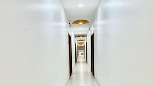 A long, narrow hallway with white walls and a tiled floor, leading to a door at the end. - Hotel Aaditya Inn Tathawade (Paud)