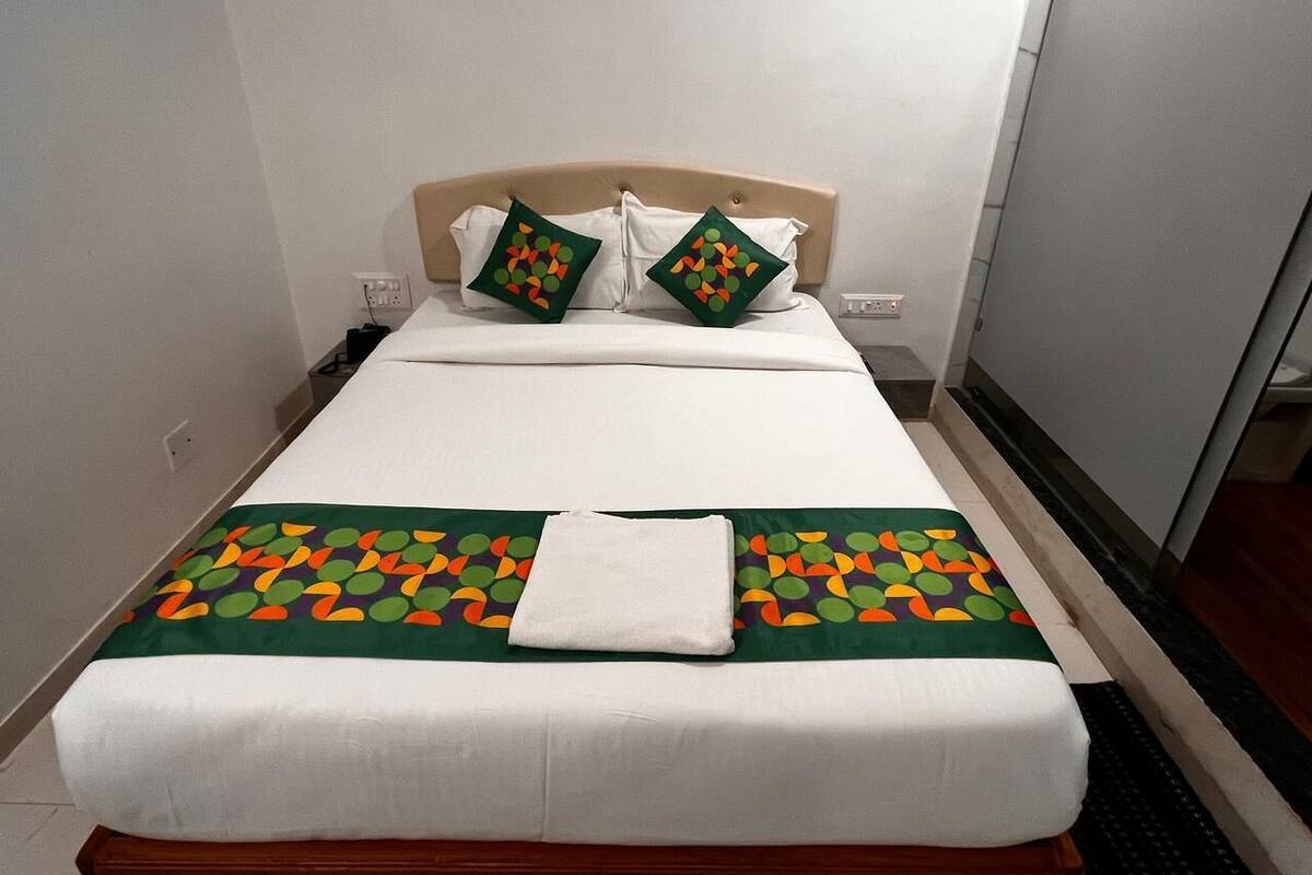 A neatly made bed with a white duvet, two pillows with a colorful geometric pattern, and a green and orange patterned bedspread.