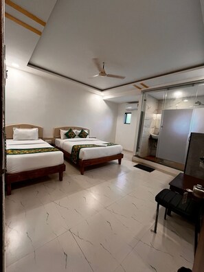 A hotel room with two beds, a ceiling fan, a small table, and a bathroom visible through a glass door. - Hotel Aaditya Inn Tathawade (Paud)