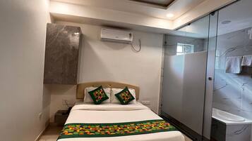 Un dormitorio con cama, ventilador de techo, equipo de aire acondicionado, mesita de noche de madera y baño con mampara de vidrio.